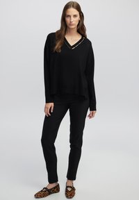 Schwarzer Strickpullover mit V-Ausschnitt und Aussparungen, kombiniert mit schwarzen eng anliegenden Hosen und Loafers im Leopardenmuster. Schlichtes, lässiges Design.