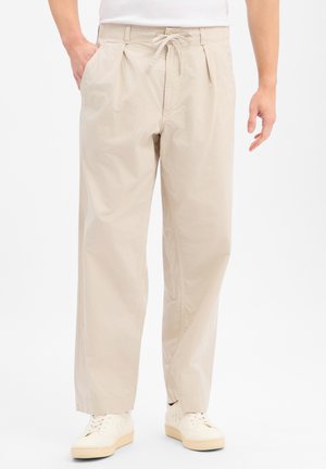 Homme portant un pantalon beige ample avec cordon et des baskets blanches, debout avec une main dans la poche sur fond blanc.