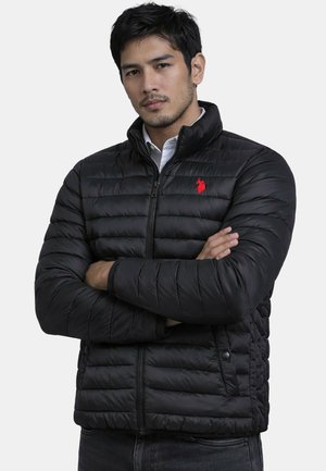 Man with folded arms wearing a black quilted jacket with a red embroidered logo and a white collared shirt underneath.