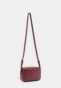 Burgundská kožená crossbody taška s obdélníkovým tvarem, zlatými detaily, texturovaným popruhem se zlatými tečkami a zipovým uzávěrem.