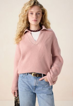 Femme aux cheveux blonds bouclés portant un pull côtelé rose, un jean bleu clair, une ceinture marron, un collier de perles foncé, et tenant un sac imprimé léopard.