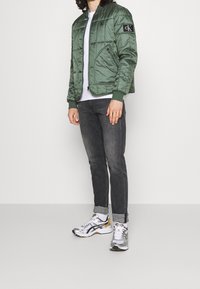 Grön vadderad jacka med ribbad krage, dragkedjsfickor och logotypapplikation, kombinerad med mörka jeans och mönstrade sneakers.