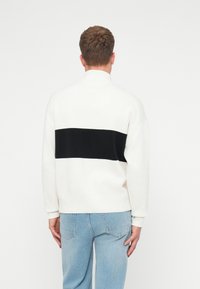 Balta džemperis ar lielu melnu horizontālu svītru aizmugurē. Mīksts audums, brīvs piegriezums, apaļš kakls, kombinēts ar gaiši ziliem džinsiem.