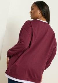 Nainen, joka käyttää löysää viininpunaista pitkähihasta paitaa valkoisen paidan päällä, katsoo sivuttain käsi lantiolla tasaisella seinällä.