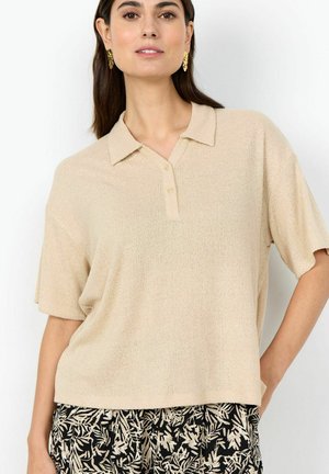 Žena v béžovom pletenom polokošeli s krátkym rukávom a čiernych nohaviciach s béžovým listovým vzorom, stojaca s jednou rukou na boku na bielom pozadí.