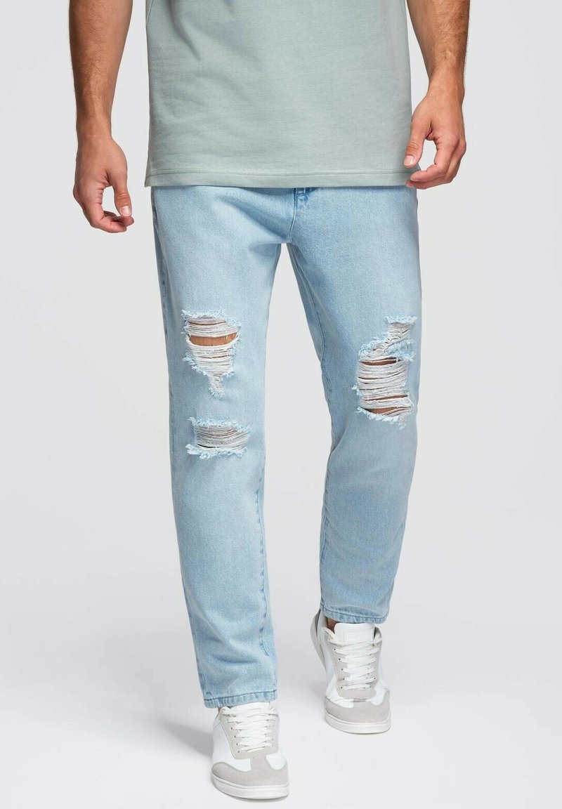 Jeans rotos de denim en azul claro, con múltiples agujeros grandes de desgaste. La emisión es recta, combinados con zapatillas blancas.