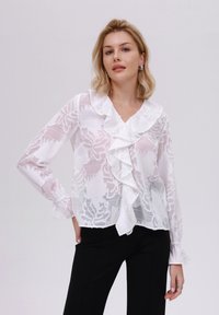 Hvit bluse med blomsterbroderi, volang i halsen og mansjettene, gjennomsiktig stoff og avslappet passform, kombinert med svarte tettsittende bukser.