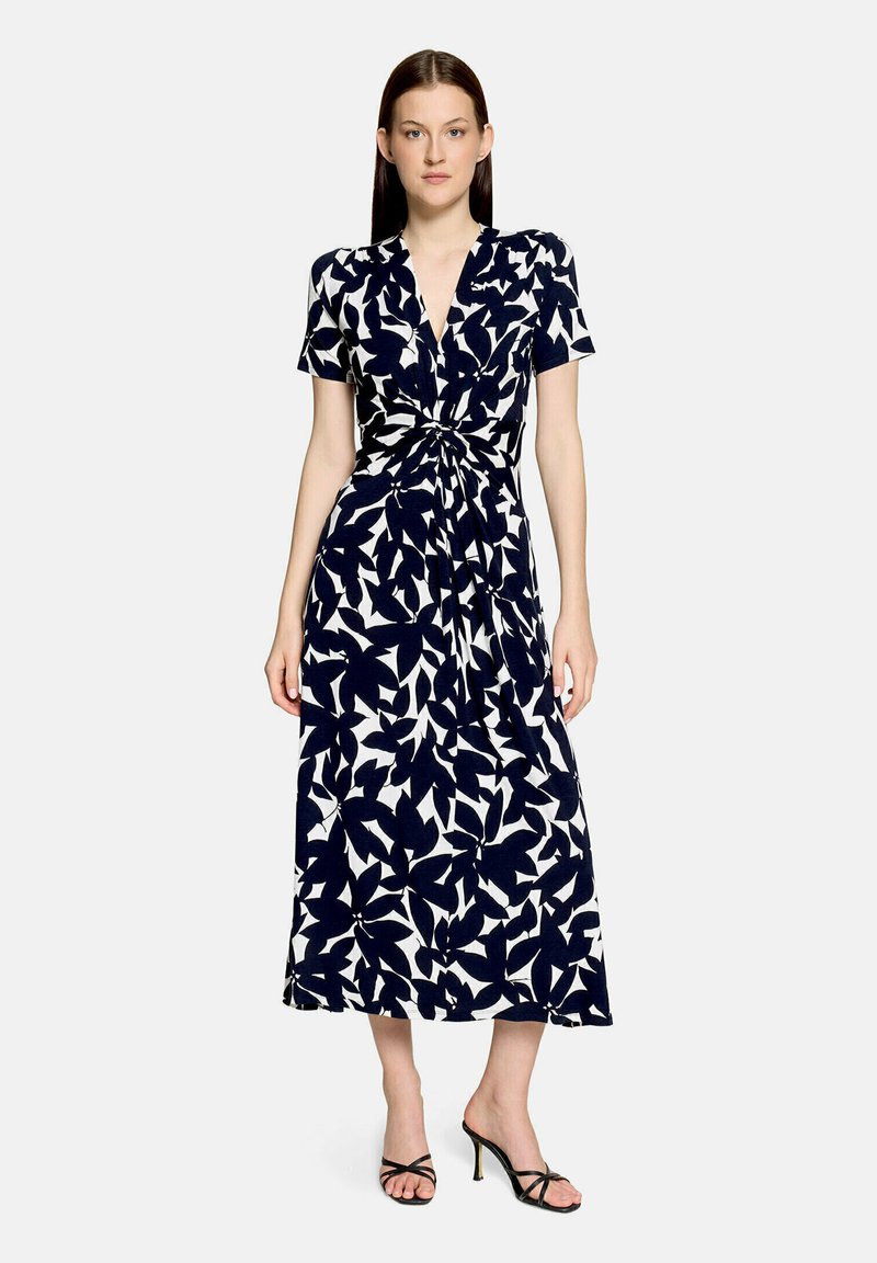 Femme portant une robe midi à motif floral noir et blanc, manches courtes et détail torsadé à l'avant, associée à des sandales noires à talons.