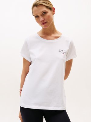 Kvinne iført en hvit Tommy Hilfiger t-skjorte med logo på brystet, stående med hendene bak ryggen mot en enkel lys bakgrunn.