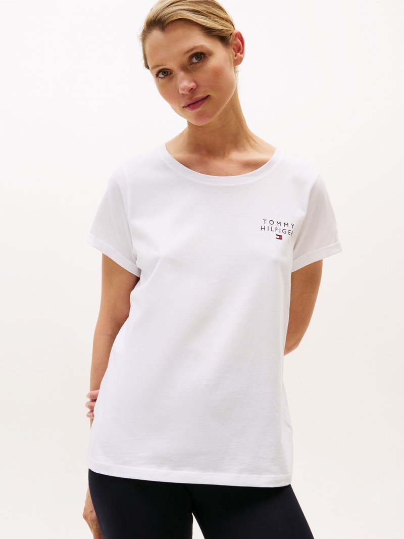 Kvinne iført en hvit Tommy Hilfiger t-skjorte med logo på brystet, stående med hendene bak ryggen mot en enkel lys bakgrunn.