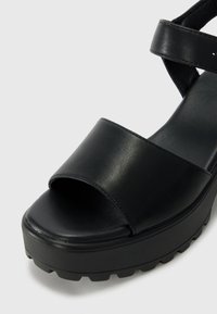 Schwarze Plateausandalen mit einem glatten Lederoberteil, offener Zehenpartie und breiten Riemen. Die Sohle ist strukturiert für besseren Halt.