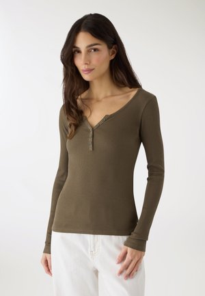 Kobieta o długich, ciemnych włosach, ubrana w oliwkowozieloną, długą, prążkowaną koszulę z guzikami, w połączeniu z białymi spodniami, stojąca na białym tle.