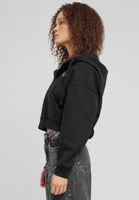 Cappuccio nero corto in tessuto morbido, con cappuccio, tasca frontale e lacci; abbinato a jeans neri sbiaditi a vita alta.