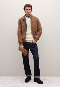 Braune Bomberjacke aus Wildleder über einem cremefarbenen Strickpullover mit Zopfmuster, kombiniert mit dunkler Denim-Jeans und braunen Handschuhen. Dunkle Schuhe runden den Look ab.