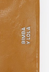 Skórzany portfel w kolorze tan z połyskiem. Na przedniej stronie znajduje się srebrny, tłoczony napis "BIMBA Y LOLA", który podkreśla gładką fakturę.