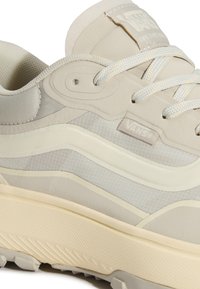 Gros plan d'une basket Vans beige avec tissu texturé, col rembourré, lacets blancs et semelle en caoutchouc épaisse et texturée.