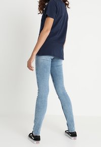 Jeans ajustados de mezclilla azul claro, con una textura suave. Combinados con una camiseta negra y zapatillas negras, vistos desde atrás.