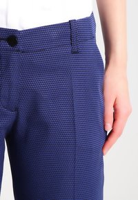 Mörkblå texturerade byxor med ett vävt mönster, med knappstängning och en hand som vilar på midjebandet för skala.