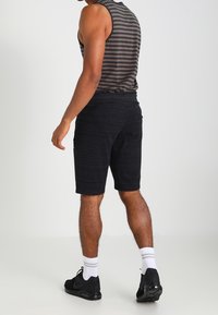 Homme portant un short de sport noir, des baskets noires, des chaussettes blanches et un débardeur gris et noir à rayures, se tenant de dos.
