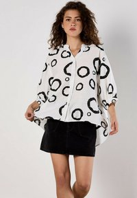 White button-up shirt with black circular patterns, loose fit, long sleeves, and a rounded hem, paired with a black denim skirt.