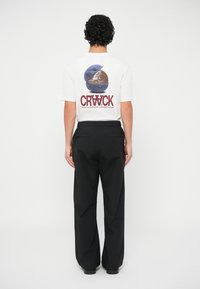 Bijela majica s okruglim grafičkim motivom na leđima u zemljanim tonovima i natpisom "CRWCK", uparen s crnim širokim hlačama.