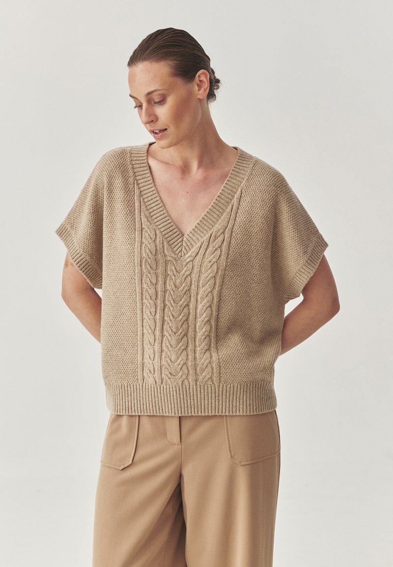 Maglione beige lavorato a maglia con scollo a V e maniche corte, caratterizzato da un motivo a trecce nella parte anteriore e orlo a coste, abbinato a pantaloni beige.