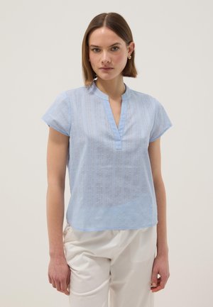 Mujer con una blusa azul claro de manga corta y textura, con escote en V y pantalones blancos, de frente contra un fondo liso.