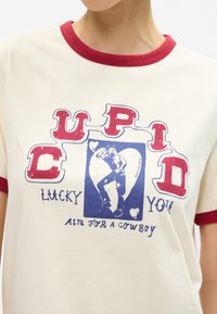 Κρεμ T-shirt με κόκκινο ριγέ κολάρο. Διαθέτει γραφικό κείμενο: "CUPID LUCKY YOU" και μια εικονογράφηση ενός καουμπόι σε μπλε χρώμα μέσα σε σχήμα καρδιάς.