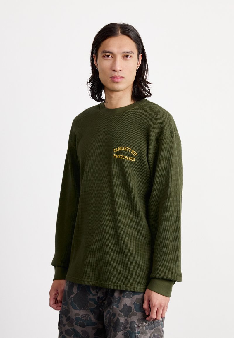 Olīvzaļš ribots garās piedurknes krekls ar apaļu kakla izgriezumu un dzeltenu izšūtu logo kreisajā krūtī. Brīvas formas.