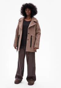 Donkerbruine wolmix jas met grote kraag, knoopsluiting en trekkoorden; gedragen over een donkere trui en gecombineerd met brede leren broeken in bruin.