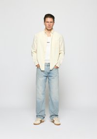 Light beige button-up shirt over a white tank top, paired with light blue denim jeans and cream sneakers with brown stripes.