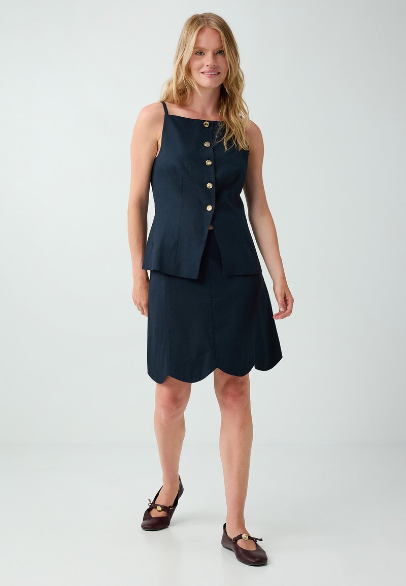 Femme vêtue d'un haut sans manches bleu marine avec des boutons dorés et d'une jupe assortie à bords festonnés, debout devant un fond uni.