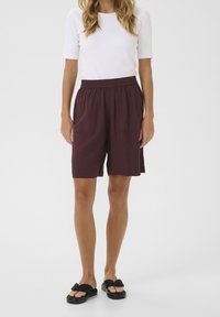 Pantaloni corti bordeaux con cintura elastica, tasche sui lati e una vestibilità rilassata, abbinati a una maglietta bianca a maniche corte e sandali neri.