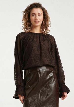 Vrouw met krullend haar die een zwarte kanten blouse met lange mouwen en een zwarte leren rok met hoge taille draagt, staand tegen een witte achtergrond.