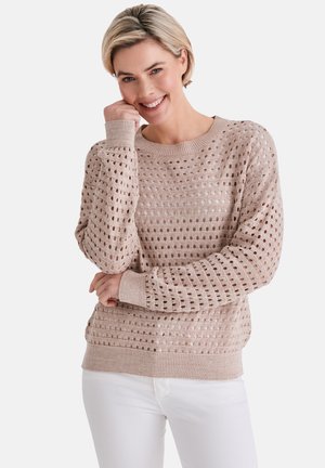 Lächelnde Frau, die einen beigen, offen gestrickten Pullover und weiße Hosen trägt, posiert mit einer Hand nahe ihrem Gesicht und der anderen, die ihre Taille kreuzt.