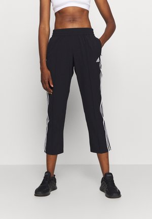 Personne athlétique portant un pantalon court noir Adidas avec des bandes blanches et des baskets noires, debout devant un fond clair uni.