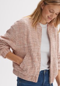 Roze textuur bomberjack met een ritssluiting aan de voorkant, gebandeerde zoom en manchetten, en zijzakken, gedragen over een wit shirt, gecombineerd met blauwe spijkerbroeken.