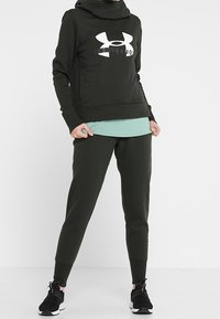 Sudadera verde oscuro y pantalones jogger a juego, hechos de una suave tela elástica. Presenta un gran logo blanco en la sudadera y un look en capas. Zapatillas negras.
