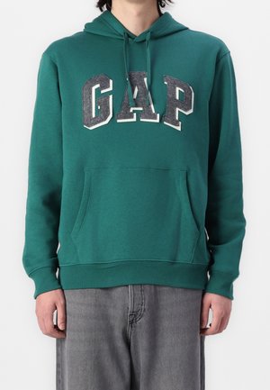 Zielona bluza z kapturem z dużymi szarymi literami „GAP” na piersi, przednią kieszenią i sznurkami, noszona z szarymi jeansami.