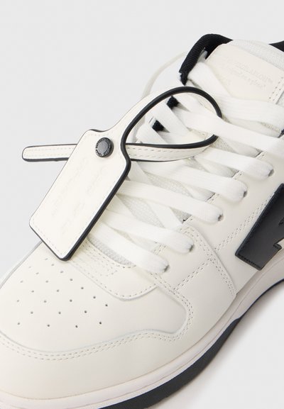 Λευκό δερμάτινο αθλητικό παπούτσι με μαύρες λεπτομέρειες, διαθέτει επίπεδες λευκές κορδόνια, τρύπες στο τμήμα των δαχτύλων και πλαστική ετικέτα προσαρτημένη στα κορδόνια.