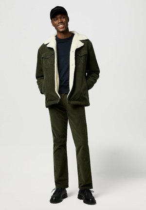 Chaqueta de algodón de color verde oliva con cuello de forro polar blanco, combinada con pantalones ajustados a juego. Unos zapatos negros completan el conjunto.