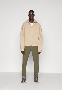 Maglione beige a zip con texture, t-shirt bianca sotto, pantaloni verde oliva e sneakers scure con suole chiare. Occhiali da sole quadrati completano il look.