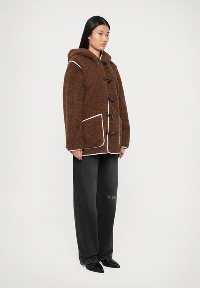 Chaqueta de forro polar marrón con capucha, ribetes blancos, cierres de madera y dos bolsillos frontales; combinada con pantalones de pierna ancha negros y zapatos negros.