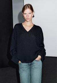 Svart stickad tröja med V-ringning, oversized passform och texturerat mönster, i kombination med ljusblå denimjeans. Enkel, modern design.