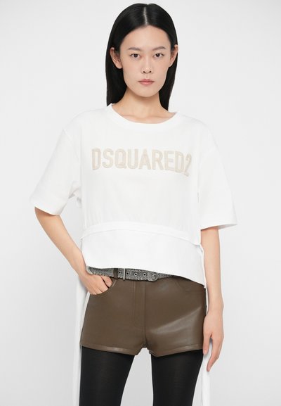 Mladá žena s dlhými čiernymi vlasmi, oblečená v bielej tričku DSQUARED2, hnedých kožených kraťasoch, čiernych pančuchách a vzorovanom opasku.