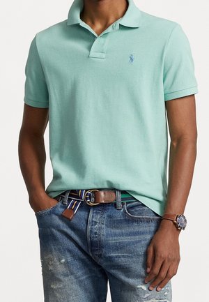 Minzgrünes Poloshirt mit klassischem Kragen, kurzen Ärmeln und einem kleinen blauen Logo. Kombiniert mit ausgewaschenen blauen Jeans und einem braunen Gürtel.