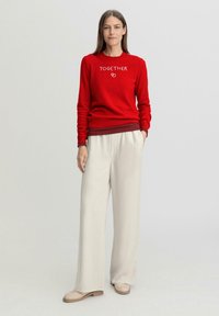 Rode sweater met de tekst "TOGETHER" in het wit, gecombineerd met lichtbeige wijde broek. Glad materiaal, casual pasvorm en minimale ontwerpaccenten.