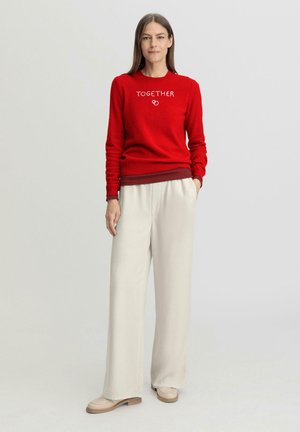 Roter Pullover mit dem Schriftzug "TOGETHER" in Weiß, kombiniert mit hellbeigen, weit geschnittenen Hosen. Glatter Stoff, lässige Passform und minimale Designakzente.