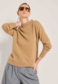 Frau in einem beigefarbenen Pullover und grauen Hosen, accessoriert mit einem großen silbernen Armband, hängenden Ohrringen und übergroßen braunen Sonnenbrillen.