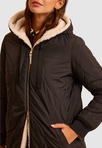 Czarna kurtka z kapturem o pikowanej fakturze, z miękką kremową podszewką, przednim zamkiem błyskawicznym i ściągaczami na rękawach. Widoczne są dwie kieszenie.